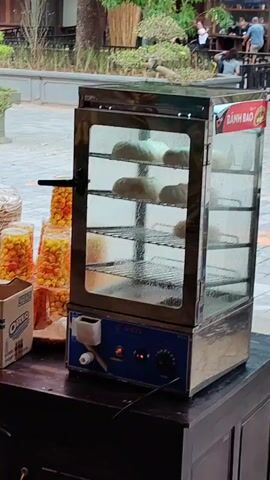 A stainless steel and glass food warmer displays steamed buns. The buns are arranged on several shelves. A box of Oreos and several containers of caramel corn are on the table next to the food warmer. People sit at tables and walk around in the background.