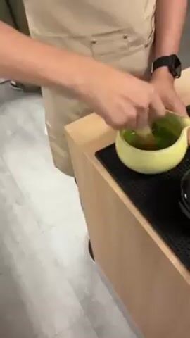 A person wearing a tan apron is whisking a green liquid in a light green bowl using a bamboo whisk on a light brown countertop. The countertop has a black non-slip mat underneath the bowl. The person is also wearing a white t-shirt and a black watch. There are two other people visible in the background, also wearing aprons. They appear to be standing at separate stations, possibly preparing similar items. The floor is a light gray tile. There is a thermostat visible on the wall.
