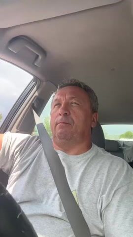 A middle-aged man with short, graying hair and a light-colored t-shirt is sitting in the driver's seat of a car. He is wearing a seatbelt. The man is looking towards the right side of the frame, then turns his head to look directly at the camera with a neutral expression. Sunlight streams into the car, illuminating his face. The interior of the car is visible, including the steering wheel, the car's roof, and the passenger seat. Outside the car, trees and a cloudy sky can be seen through the windows.