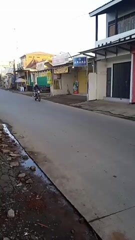 The video shows a street scene during daytime. A line of motorcycles is driving down a gray asphalt road. Buildings and small businesses line the sides of the street, most are one or two stories tall. The sky is light blue. There is some loose stone and dirt on the roadside. The road is mostly clear except for a few puddles. A cell tower is visible in the distance. Two motorcycles pass by the camera.
