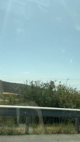 The video shows a view from inside a moving car, looking out the side window. The car is driving on a highway with a guardrail along the side of the road. The landscape consists of dry, scrubby vegetation and hills. In the distance, there are some agricultural fields and structures. As the car moves, the view changes to reveal a billboard and a bridge overpass. The sky is blue with some haze. The car's side mirror is visible in the lower right corner of the frame.