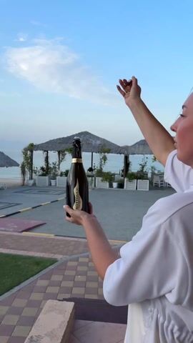 A woman in a white shirt holds a bottle of Bottega Prosecco DOC Vintage. The camera focuses on the bottle in her hand, with a beach, the ocean, and a blue sky with some white clouds in the background. In the background, there are several straw-roofed cabanas. The woman is facing away from the camera, and looking out at the scene in front of her. The ground is brick and cement. There is grass to the side.