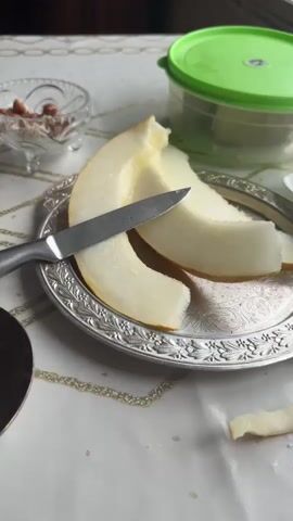 A close-up shot shows slices of melon on a decorative silver plate. A knife rests on the melon. To the left, a glass of dark tea sits on a saucer. A hand enters the frame and picks up the tea glass. In the background, a small glass bowl holds nuts and a green-lidded container. The scene is set on a white tablecloth with a gold pattern.