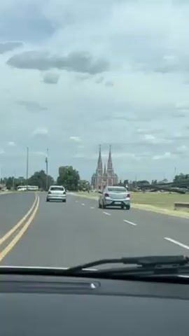 The view from inside a car driving down a highway. The focus is on the road ahead, where two silver cars are traveling in the same direction. In the distance, a large church with two tall spires is visible. The sky is partly cloudy. There are yellow lines on the highway. The car’s windshield wipers are visible at the bottom of the frame. Music is playing in the background.
