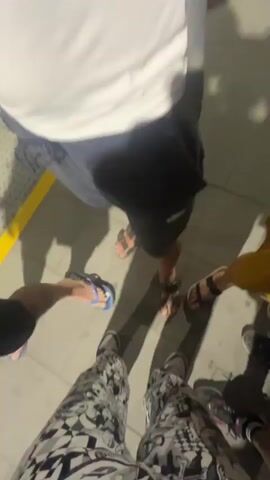 The video begins with a POV shot looking down at several pairs of feet wearing various types of footwear, including sandals, sneakers, and flip-flops. The ground appears to be paved. Shadows are cast on the ground, indicating a light source. The camera then pans up and out to show a nighttime scene with streetlights illuminating palm trees and a fence. The overall impression is of a group of people standing together outdoors at night.