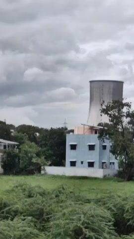 A video captured from a moving vehicle shows a large, conical cooling tower against a cloudy sky. The tower is surrounded by a cluster of low-rise buildings and trees. The foreground consists of a grassy field, some of which appears wet. The video begins with a blurred view of lush green foliage and a large tree trunk, then transitions to the clearer scene of the cooling tower and surrounding landscape.