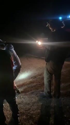 The video begins at night with two figures in the foreground, facing away from the camera. A red vehicle with its headlights on is visible in the background. The vehicle has "SPASIORI" and "GASCI" written on its side, and a crest on the door. In the second frame, the figures are more illuminated, and a person in the foreground is wearing a headlamp. The third and fourth frames show the same scene with more people visible around the vehicle. The lighting in the scene is from the vehicle's headlights and at least one headlamp. The ground appears to be covered in grass and dirt. The overall environment appears to be outdoors in a rural or undeveloped area.