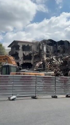 The video shows the demolition of a multi-story building. An excavator with a hydraulic arm is actively breaking down the structure, causing dust and debris to fly. In the foreground, a metal barricade is erected, with signs indicating "MERT GRUP YIKIM HAFRIYAT" and phone numbers. Two construction vehicles, likely excavators, are parked behind the barricade. The sky is partly cloudy. To the right, a green tree is visible, and a sign with a "no right turn" symbol and text is present. The overall scene depicts a construction site where demolition work is underway.