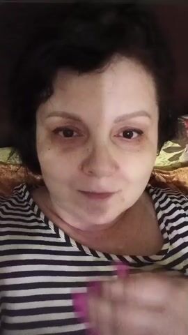 A woman with dark hair and dark circles under her eyes, wearing a black and white striped shirt, blows a kiss. Her hand, adorned with long, bright pink fingernails, covers her mouth for a moment before moving away. The background appears to be indoors, with hints of a patterned blanket or bedding.