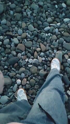 The video shows a person in jeans and sneakers standing on a rocky beach. The camera then pans up to show a young child in a yellow jacket and baseball cap walking towards the sea at sunset.