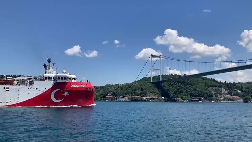 A large white and red ship, bearing the Turkish flag and the name "ORUÇ REİS" on its side, sails on a body of blue water under a bright, cloudy sky. The ship is moving from left to right across the frame. In the background, a large suspension bridge crosses the water, with lush green hills and buildings visible on the far bank. The bridge is a prominent feature, stretching across the scene. The water is calm, with gentle ripples visible. The overall scene is set during daylight with clear weather.