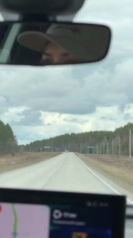 The video captures a view from inside a car, looking out through the windshield onto a straight, rural road. A digital map display is visible in the foreground, showing a green route. The road stretches out towards the horizon under a sky filled with gray, cloudy patterns. A line of green trees borders both sides of the road. A couple of road signs can be seen on the right side of the road, one bearing text in a language other than English. In the car's rearview mirror, the reflection of the driver is visible, wearing a light-colored hat. Another car is also visible in the distance on the road ahead.