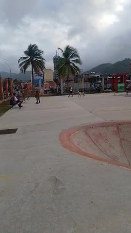 A young boy wearing a yellow shirt, blue jeans, a helmet, and black shoes is skateboarding in a concrete skatepark on a cloudy day. He skates around a bowl-shaped feature with red and blue edges, rides over a bump, and maneuvers near a metal grate. The skatepark contains other obstacles such as a low flat bar. In the background, there are palm trees, buildings, mountains, a fenced-off area, and people walking around. The area is paved with concrete and has some cracks. Other structures within the park include benches and decorative fencing.