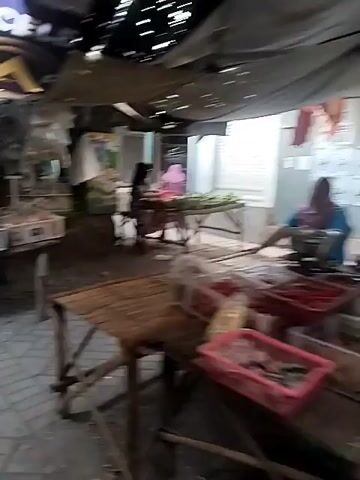 A shaky, blurry video shows a woman walking through a market stall. She is wearing a light pink hijab, a grey long-sleeved shirt, and patterned green pants. She carries a white plastic bag in her right hand and has a small brown bag slung across her chest. Behind her, a market stall is visible with various items on display, including what appears to be food items. The stall has a wooden structure with a tarp overhead. The ground is paved with interlocking bricks. Further into the market, other people and stalls are faintly visible.