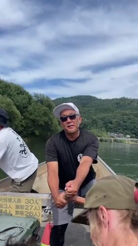 A man in a black t-shirt, grey shorts, a white baseball cap, and sunglasses is sitting in a boat and rowing with a wooden oar. He is in the foreground of the video. Behind him, another man in a white t-shirt with a black wave design and a black hat is standing and also rowing with a wooden oar. The boat is on a body of water, with green, tree-covered hills in the background. The sky is cloudy. There is some text visible on the side of the boat. A portion of a person with blonde hair and a green hat is visible in the lower right corner of the frame.