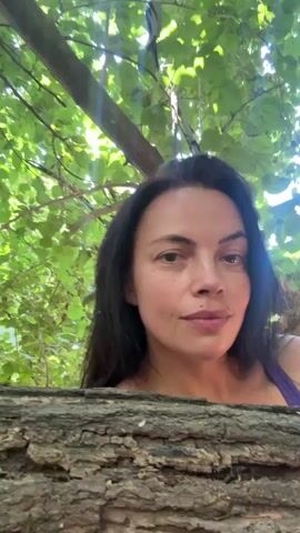 A close-up, head-and-shoulders shot of a woman with dark hair and light skin is captured from a low angle. She is positioned behind a thick, horizontal log of wood. The woman is looking directly at the camera with a neutral expression. Her hair is dark and falls around her face. She is wearing a purple top. The background is filled with lush green leaves and branches of trees, suggesting an outdoor, natural environment. Sunlight filters through the foliage, creating dappled light on the scene.