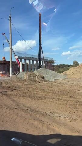 The video captures an outdoor construction site on a sunny day with a clear blue sky and scattered clouds. The foreground shows a dirt area with tire tracks, indicating vehicle activity. In the midground, a large pile of gravel is visible, along with construction materials and equipment. A prominent feature is a tall, dark construction crane with a long boom, positioned near a concrete structure under construction. To the right of this crane, a tall, slender construction tower, possibly a pile driver or a concrete pump, stands vertically. Behind this tower, a bridge-like structure is being built, supported by multiple concrete pillars. Further in the background, on the left, another crane is actively lifting a red object, likely a bucket or container. The overall scene depicts an active construction project, possibly for a bridge or overpass, with various heavy machinery and materials on-site. There are no identifiable people clearly visible in the frame.