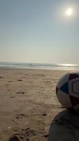 The video shows a beach on a sunny day. A soccer ball rests in the foreground on the sand. In the distance, two people are in the ocean. The sky is clear and bright. The sun is high in the sky.