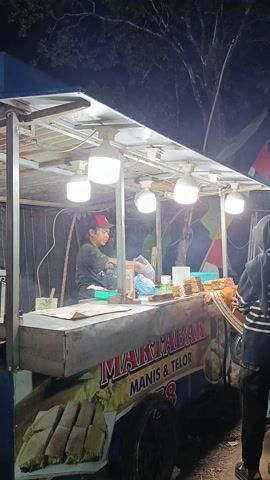 This media file depicts a nighttime scene at an outdoor market or fair in Sambirata, Indonesia. The primary subjects are a group of people, likely vendors and customers, gathered around food stalls. One stall prominently features a banner with the text "MARTABAK MANIS & TELOR" in large, colorful letters, indicating the sale of sweet and savory Indonesian pancakes. The stall is illuminated by bright overhead lights, highlighting trays of prepared food and various ingredients.

Several individuals are visible, including a vendor wearing a black jacket and a red cap, seemingly attending to customers or preparing food. Other people, dressed in a mix of casual and traditional attire, are browsing or waiting in line. The overall atmosphere suggests a lively, community-oriented event. Colorful flags and banners are strung up, adding to the festive ambiance. The background shows trees and dark foliage, consistent with an outdoor setting at night. The ground appears to be paved or made of dirt. The lighting suggests it is nighttime, with artificial lights from the stalls being the main source of illumination. The weather appears clear. The presence of multiple food vendors and the gathering of people suggest a market or festival, possibly a night market.