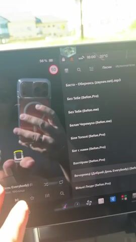 A person's hands are visible on the left side of the screen, interacting with a car's touchscreen display. The screen shows a list of songs with Ukrainian titles and artist names, as well as a parking assist feature with a car icon. The car appears to be in park. The background shows a blurred outdoor scene with buildings and trees. The time is 16:00 and the temperature is 32 degrees Celsius. The screen also displays battery percentage and speed limit information.