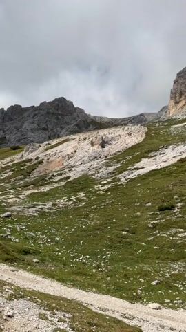 The video pans across a mountainous landscape with green grass and rocky slopes under a cloudy sky. A dirt path is visible in the foreground, and a trail marker with red arrows points upwards. Two small figures, likely hikers, are visible on the slope in the distance.