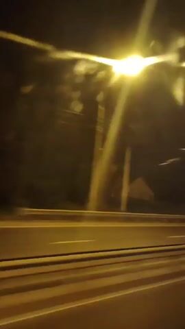 This is a short video clip showing a view from a moving vehicle at night. The scene depicts a highway with multiple lanes, indicated by yellow and white lines on the asphalt. The road appears wet, suggesting recent rain or damp conditions. On the side of the highway, there are trees and some structures that are blurred due to the motion. A lamppost illuminates the road with a bright light, creating streaks in the video. Other vehicles are also visible as streaks of light, indicating they are moving at high speed. The overall atmosphere is dark and fast-paced, characteristic of nighttime travel on a highway. There is no discernible text in the video. The location is contextually identified as Zavidovo, Russia.