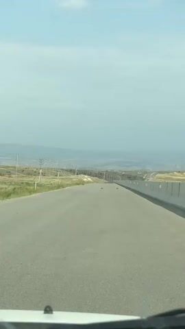The video captures a first-person perspective of driving on a paved road. The road appears to be a highway with multiple lanes, though only one lane is clearly visible in the frame. On the left side of the road, there is a grassy verge with scattered telephone poles. On the right side, there is a concrete barrier. In the distance, the road curves and ascends a gentle slope, leading towards a hazy horizon with mountains. The sky is overcast with some clouds. The vehicle's dashboard is partially visible in the foreground. The overall environment appears to be rural or semi-arid.