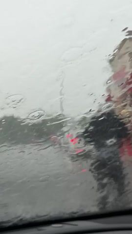 From inside a car, the windshield is covered in rain.  Visibility outside is low.  Red tail lights of a car ahead are visible through the rain.  Rain streams down the windshield, obscuring details of the surroundings.