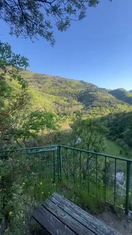 The video shows a scenic view from a vantage point overlooking a valley. The valley is lush and green with trees covering the hillsides. A green metal railing and a wooden bench are in the foreground. The sky is clear and blue. The scene appears peaceful and natural. There are some buildings visible in the valley.