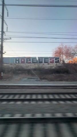 The video shows a view from a moving train. The focus is on the railroad tracks, which are made of gray metal and have a textured surface. Alongside the tracks, there's a patch of land with debris and some vegetation. In the background, there's a large brick building with graffiti, including the word "HOPELESS" in large block letters. Power lines and supporting structures are visible above. The sky is a pale blue. The camera moves with the train, blurring the foreground details while keeping the background somewhat steady.