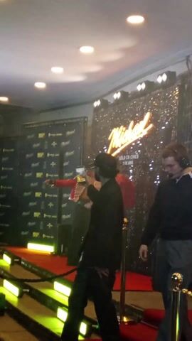 Here is a description of the video:

The video shows a red carpet event for the film "Michael." In the background, there is a sign that reads "Michael, solo en cines, 13 de abril." Someone is standing on the red carpet dressed in an outfit similar to what Michael Jackson wore. As the person in the costume points and gestures towards the camera, a burst of sparks shoot up, creating a brief, dramatic effect. People stand around watching the performance.