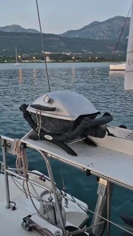 A close-up shot focuses on a silver Weber grill with black legs, positioned on a white shelf attached to the railing of a sailboat. The grill has a built-in thermometer on its lid. The boat is on the water, and in the background, several sailboats are anchored. Beyond the water, there are mountains and a coastline with lights from buildings. The sky is a soft, pale blue, suggesting it is either dawn or dusk. The water is rippling gently, and there are reflections of the boats and the sky on its surface. Ropes are visible on the boat's deck, securing it. The scene is calm and scenic, with a focus on the grill as a potential element for an outdoor meal on the water.