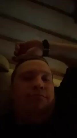 A man with fair skin and short hair is lying down and looking at the camera. His arm, adorned with a black smartwatch, rests on his forehead. He is wearing a black shirt. The background shows a textured ceiling and muted green lighting.