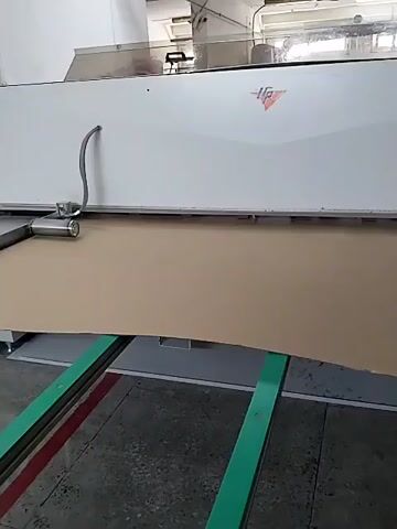 A close-up, static shot shows a large sheet of tan material being fed into a white industrial machine. The material is supported by two green conveyor belts. A metal nozzle attached to a cable extends from the left side of the machine, hovering just above the tan material. The machine has a logo in the upper right corner featuring red triangular shapes and the letters "VP". The environment appears to be a factory or workshop with a concrete floor. The video is 4 seconds long and shows the material moving slowly into the machine.
