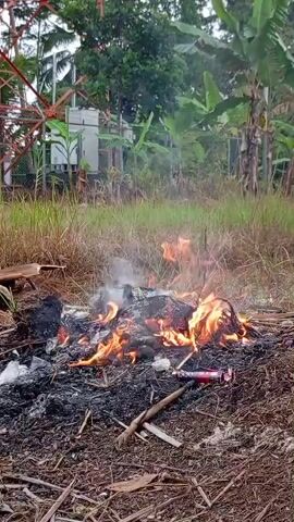 The video shows a small fire burning in an outdoor area. The fire is consuming various types of debris, including what appears to be paper, plastic, and possibly some organic material. Flames are visible, with smoke rising from the burning pile. In the background, there is tall grass and vegetation, suggesting a rural or semi-rural setting. A large metal tower structure, possibly a communication tower, is also visible in the distance, along with some equipment housed in a small building. The ground around the fire is covered in dirt, leaves, and twigs. The overall impression is of an uncontrolled burn of trash or yard waste.