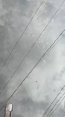 The video shows a grey, textured surface with several dark, thin lines running diagonally across it. These lines appear to be wires or cracks. In the bottom left corner, a small, weathered object, possibly made of wood and metal, is visible. The surface is speckled with small white particles, creating a slightly grainy texture. The lighting is diffuse, casting a muted tone over the scene.
