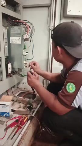 A person wearing a black cap and a brown and gray uniform is kneeling and working on electrical wiring inside a control panel. The person is holding a wire in their left hand and a tool, possibly wire strippers or pliers, in their right hand, and is actively engaged in connecting or disconnecting a wire. In front of the person, on the floor, are several tools including a pair of red-handled wire cutters, a yellow-handled tool, and a multimeter. A cardboard box labeled "OHNT" is also visible. The control panel contains various electrical components, including a large silver unit with wires connected to it and a smaller black component with wires. The environment appears to be an indoor industrial or maintenance area with a wooden or metal floor.