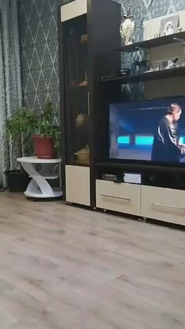 The video shows a living room with wood-patterned flooring and a patterned wallpaper. On the right side of the frame, there is a large dark brown and cream-colored entertainment unit. A television is mounted in the center of the unit, displaying a scene with four people. A plant in a red pot is on a small white table to the left of the entertainment unit. The lighting appears to be artificial, and the scene is stationary.
