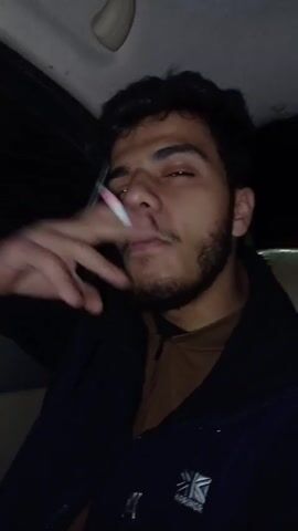 A young man is sitting in the driver's seat of a car, holding a lit cigarette between his fingers. He is wearing a brown zip-up shirt under a black jacket with the "Karrimor" logo visible on the right side. He has curly hair and a trimmed beard. He exhales smoke from his mouth and smiles slightly. The car's interior is dark, with a beige headliner visible above. The scene appears to be at night.