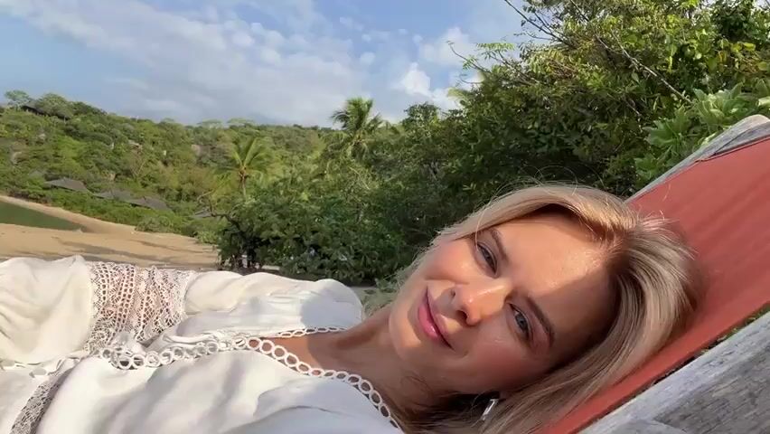 The video opens with a close-up of a fair-skinned woman with blonde hair smiling directly at the camera. She is lying down, wearing a white garment with lace detailing. The background shows a sandy beach, calm blue water, lush green hills, and a bright sky with scattered clouds. As the camera pans away, the perspective shifts to show the woman's legs, still resting, and the beach environment more broadly. We see the shoreline, the water with a few boats in the distance, and the hilly, tree-covered landscape. The shot continues to pan, offering wider views of the bay and the horizon with more hills. The video concludes with a view of the woman's legs again, this time from a slightly different angle, with the beach and ocean as the backdrop. The scene appears to be a tropical or subtropical beach resort on a clear day.