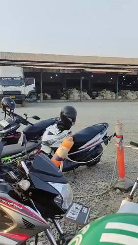 The video captures a parking area filled with numerous motorcycles, suggesting a bustling location, possibly a market or a logistics hub, given the presence of a large warehouse structure and several trucks in the background. The ground is covered in gravel, typical of outdoor parking lots. Several motorcycles are parked closely together, some with helmets placed on their seats or handlebars. In the mid-ground, a few individuals are visible near some of the motorcycles, though their actions are indistinct. The lighting suggests it is either early morning or late afternoon. A tall power line tower is visible in the far background, indicating the presence of electrical infrastructure nearby.