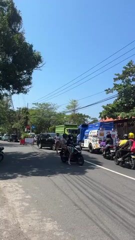 The video shows a busy street scene in what appears to be a tropical or sub-tropical location, with bright sunlight and a clear blue sky. The perspective is from a person riding a white scooter, and the camera captures the road, passing vehicles, and buildings along the side.

In the foreground, the handlebars and part of the instrument panel of the scooter are visible, along with a side mirror. The side mirror reflects the rider's face, wearing a helmet and a dark shirt. A red string is tied to the mirror mount.

On the road, several vehicles are moving. A white scooter with two riders, wearing helmets and casual clothing, is in front of the viewer's scooter. To the right of this scooter, another scooter with a single rider wearing a yellow helmet is visible. Further ahead, a blue and white truck is driving. Behind this truck, a white van is also present. To the far left, a dark-colored car is seen. On the right side of the road, a silver car is driving, and further back, another white car is visible.

Buildings with red-tiled roofs line the right side of the street. Some of these buildings appear to be shops or businesses, with awnings and visible interiors. Trees and greenery are present, suggesting a well-vegetated area. Power lines are strung across the sky.

The overall impression is of a lively street during the daytime, with typical road traffic and the presence of local architecture.