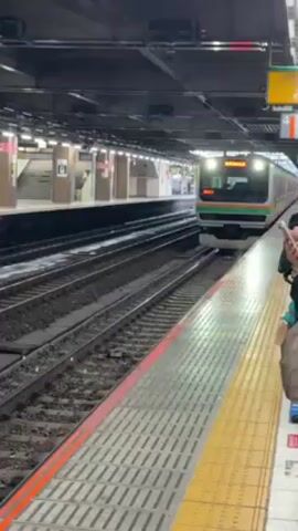 The video shows a train approaching a platform at a train station. The train is painted with silver and green stripes, and has a display showing the name of its destination. There is at least one person standing on the platform near the edge, holding a phone, seemingly recording the train as it approaches. The station appears to be indoors, with a tiled platform and overhead infrastructure.