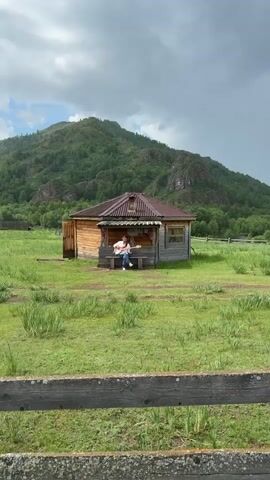 A person sits on a bench in front of a wooden house and plays a guitar. The house has a porch with a corrugated metal roof. The person is wearing a white shirt and blue jeans. The house is surrounded by a grassy field, and in the background, there is a large, tree-covered mountain under a cloudy sky. A wooden fence is visible in the foreground.