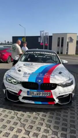 The video shows a white BMW with a distinctive racing livery parked outdoors on a paved surface. The car features a color scheme of white with blue, red, and black stripes, as well as numerous sponsor logos and the words "Safety Car" across the hood and roof. The license plate reads "BM WP 9991." Three men are standing around the car, seemingly conversing, with one man handing something to another. In the background, a red Tesla is visible, along with buildings and flags bearing logos. The sky is clear, suggesting a sunny day.