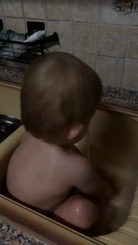 A baby sits in a sink, playing with water from a faucet. The baby is facing away from the camera, with their back to the viewer. They have light brown hair and are a light-skinned child. The baby's hands are wet, and they appear to be splashing the water around. The sink is a light brown color, and the surrounding area is tiled with beige squares. The lighting suggests it might be daytime indoors. There is no visible text in the image.