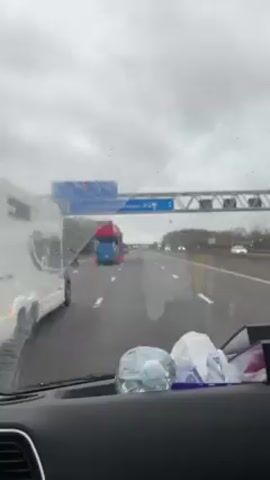 The video shows a car driving on a highway on a cloudy day. The windshield has raindrops on it, and the wiper is intermittently in use. A large, blue car carrier truck, carrying several cars, is ahead in the same lane. Other vehicles are visible further down the highway. The dashboard of the car is visible, with a water bottle, a tissue, and a package of wet wipes. The video is shaky due to the movement of the car.