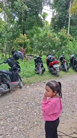 The video features two young children standing on a gravel path in an outdoor setting. A young girl, wearing a pink long-sleeved top and black pants, stands with her back partially to the camera, looking to her left. Her hair is tied back in a ponytail. Next to her, a young boy in a grey t-shirt with a graphic (partially visible, resembling a camera icon and text like "Screa") and blue ripped jeans stands with his hand to his ear, initially looking forward then turning towards the background.

Behind the children, several scooters and motorbikes are parked off the path on a grassy area. Beyond the parked vehicles, dense tropical vegetation, including palm trees, forms a lush green backdrop. A few adults are visible further in the background amidst the motorbikes and trees. The scene suggests a daytime gathering, possibly in a rural or village area in Meunasahreudeup, Indonesia, given the natural surroundings and typical local transport. The children appear pensive or observant. A black cable lies on the gravel path near their feet.