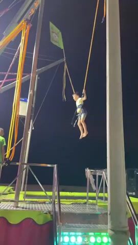 The video shows a child on a bungee trampoline ride at night. The child is wearing a harness and is attached to bungee cords that are connected to an overhead structure. The child is in mid-air, having been launched upwards by the trampoline. In the background, there is a man standing on a platform and a dark sky. The ride appears to be at an amusement park or fair, as indicated by the colorful lights and the general atmosphere. The child is alone on the trampoline at this moment.