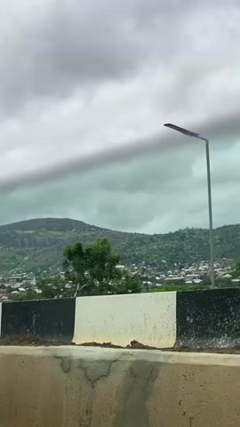 The video shows a landscape with a large mountain in the background. The sky is cloudy and overcast. In the foreground, there is a road with a barrier. There are streetlights visible along the road. A van is seen driving on the road. The van has text on its rear window. The scene appears to be a town or city nestled at the foot of the mountain. The overall atmosphere is somewhat muted due to the cloudy weather.